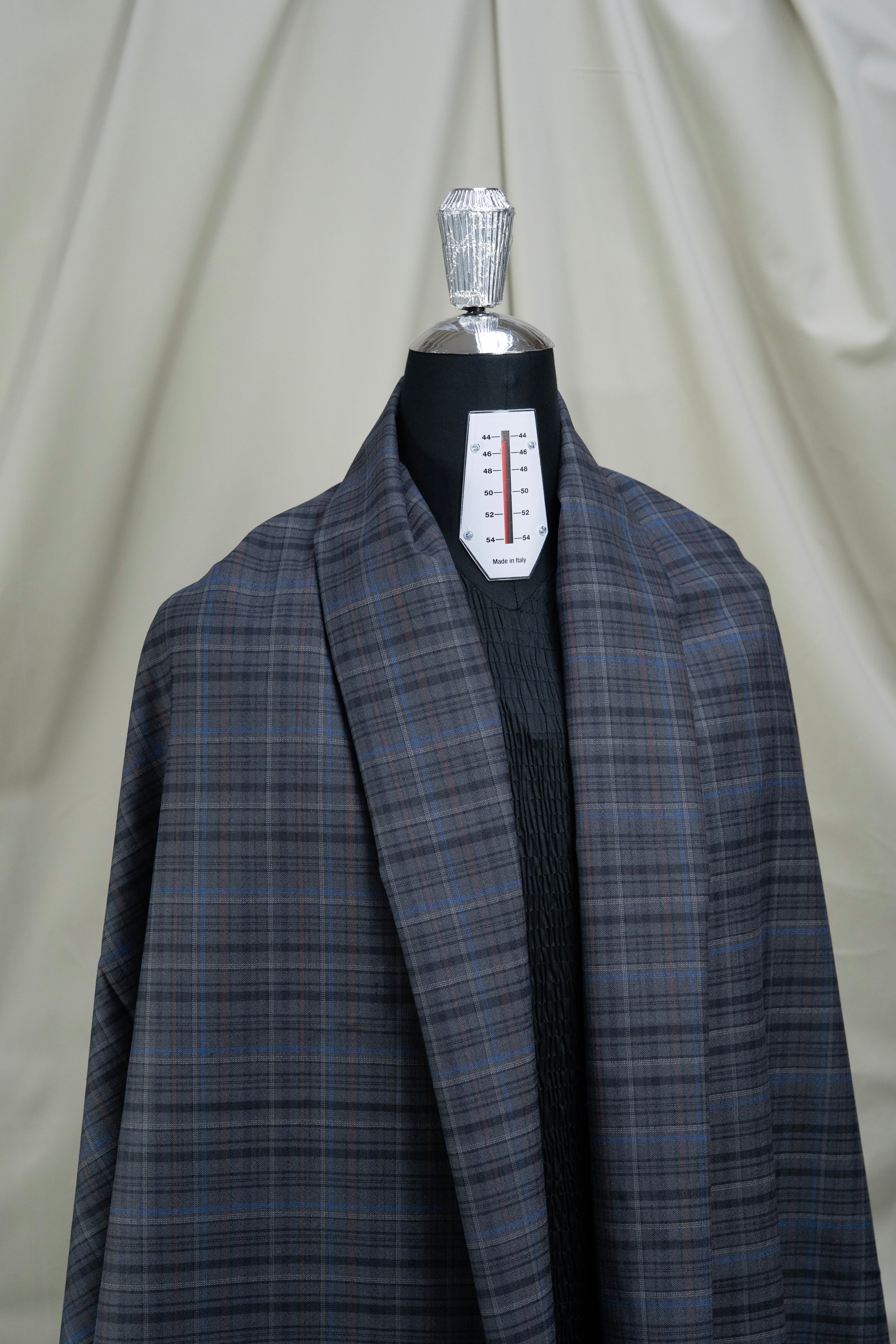 A dark gray and blue plaid coat displayed on a black mannequin torso with a silver neck cap, set against a draped beige fabric backdrop.